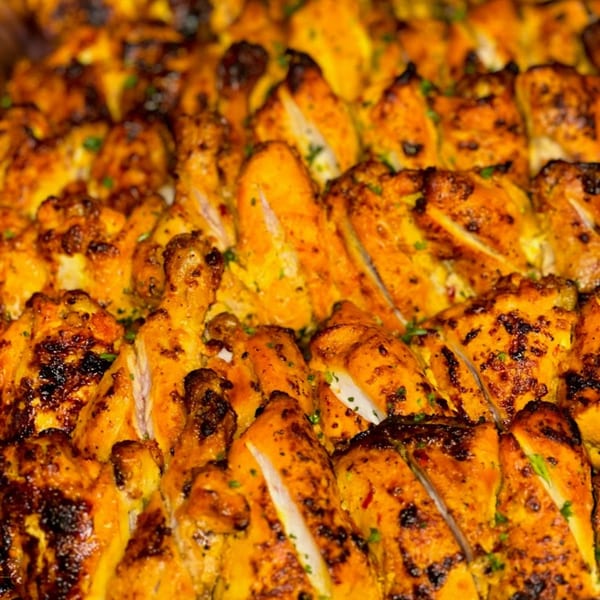 Close-up of golden-brown grilled chicken pieces, some sliced, with charred edges and green herbs.