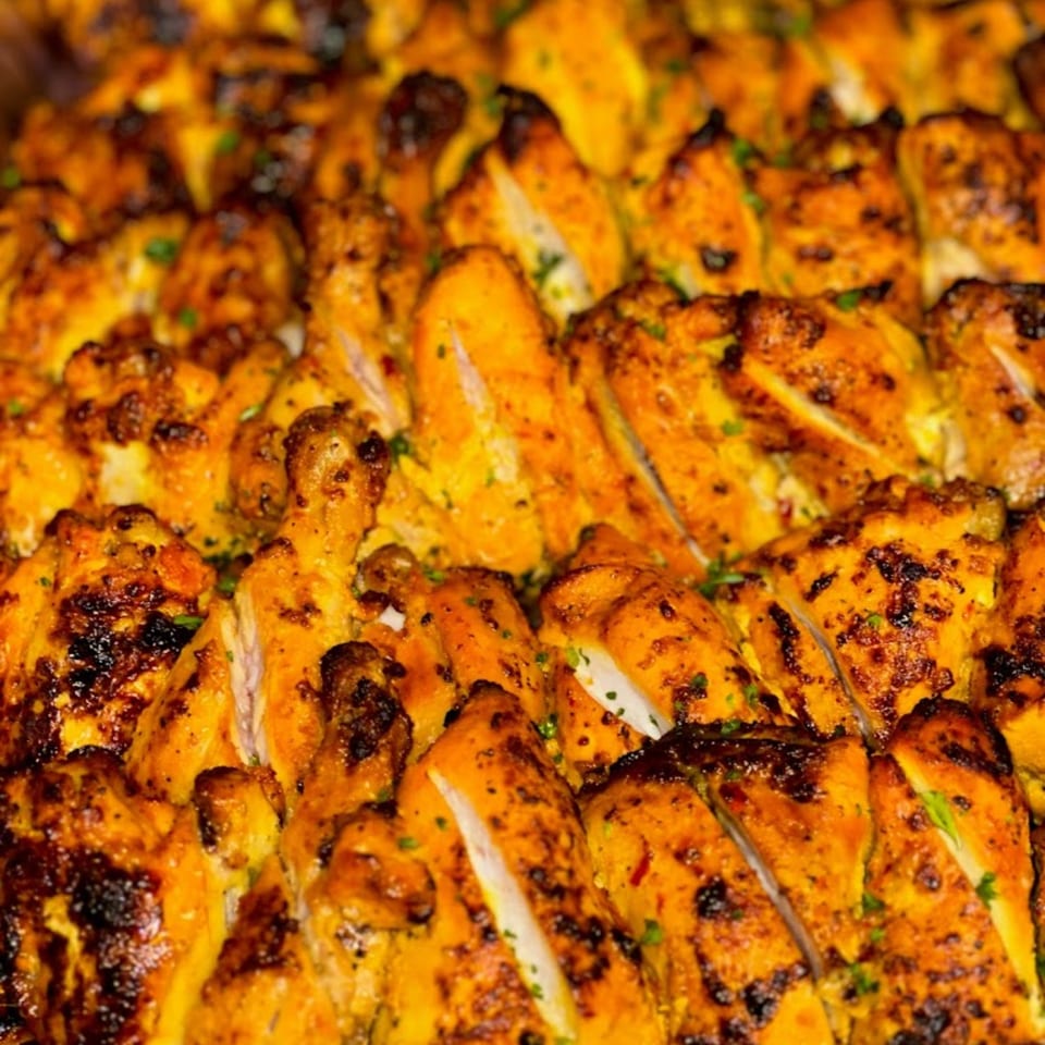 Close-up of golden-brown grilled chicken pieces, some sliced, with charred edges and green herbs.