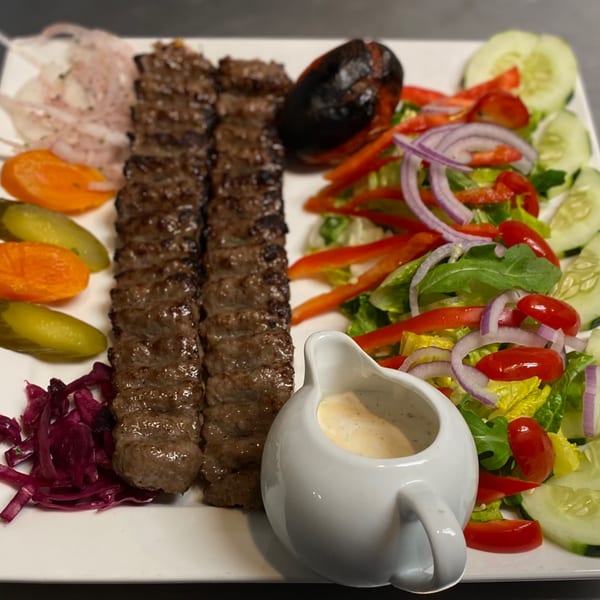 Two skewers of Persian koobideh kebab with grilled tomato, salad, pickled vegetables, and sauce.
