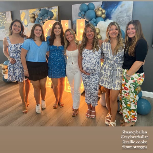 Seven women smiling, posing together in a room decorated with blue and gold balloons.