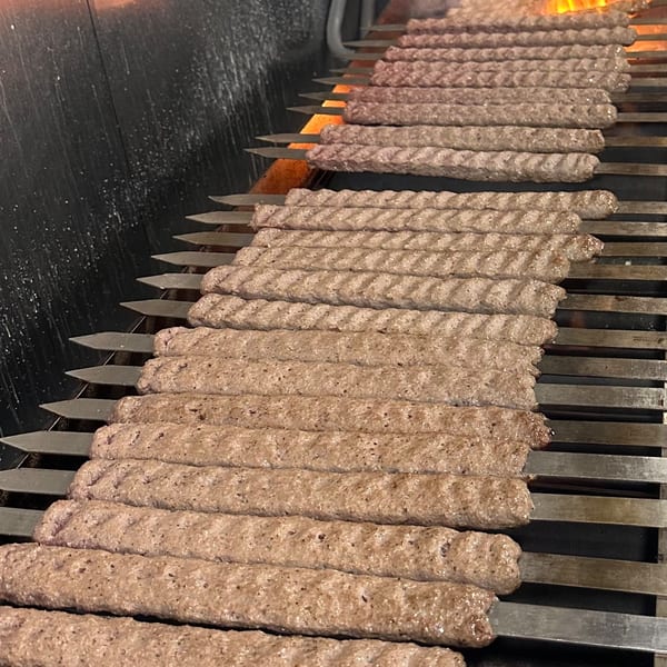 Rows of Persian koobideh kebabs cooking on a grill with flames visible in the background.