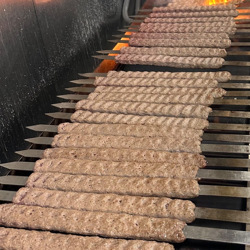 Rows of Persian koobideh kebabs cooking on a grill with flames visible in the background.