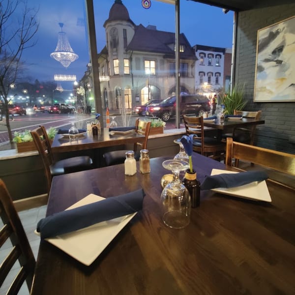 Restaurant interior with set tables and a large window overlooking a street at dusk.